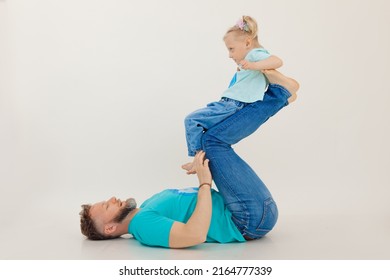 Young Athletic Man With Pumped Up Arms Lying On Back Holding And Lifting Up With Legs Little Girl, White Background. Whole Family Workout, Father And Daughter. Sport And Healthy Lifestyle Concept