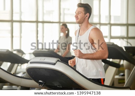 Young athletic man running on treadmill. Handsome muscular guy working out at gym and listening to music with headphones. Enjoy your lifestyle.