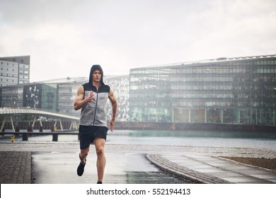 Young Athletic Man In Sportswear Running In Rain Down Street. 