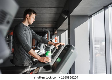 Young athletic man standing on treadmill in gym. In one hand he holds a green water bottle and the other touches the treadmill screen. - Powered by Shutterstock