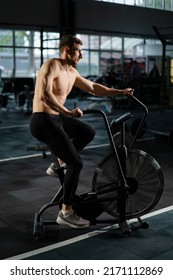 Young Athletic Middle Aged Man Training On Air Resistance Bike, Cross Training Workout Set In Gym. Active Man Spinning A Air Bike In Gym With Trainers. Male Training On Air Bike.