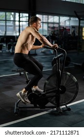 Young Athletic Middle Aged Man Training On Air Resistance Bike, Cross Training Workout Set In Gym. Active Man Spinning A Air Bike In Gym With Trainers. Male Training On Air Bike.