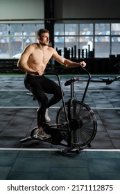 Young Athletic Middle Aged Man Training On Air Resistance Bike, Cross Training Workout Set In Gym. Active Man Spinning A Air Bike In Gym With Trainers. Male Training On Air Bike.