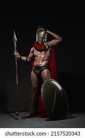 A young athletic sexy man dressed as a Roman warrior in a red cloak stands with a spear in his hand and a full-length shield on a black background.