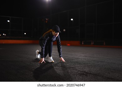 Young Athletic Woman In Black Tracksuit And Cap Is Standing In The Low Start Position Before Running, Jogging At City Night Alone, Outdoors.Sports Healthy Lifestyle. Side View Portrait. Copy Space