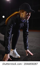 Young Athletic Woman In Black Tracksuit And Cap Is Standing In The Low Start Position Before Running, Jogging At City Night Alone, Outdoors.Sports Healthy Lifestyle. Side View Portrait. Copy Space