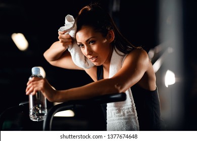 Young Athletic Woman Feeling Exhausted While Cycling On Exercise Bike And Wiping Sweat With A Towel In Fitness Center. 