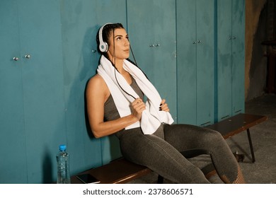 Young athletic woman sitting on a bench with a towel, headphones and listening to music in the locker room - Powered by Shutterstock