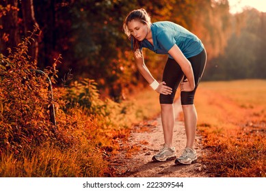 Young athletic woman taking a break from training standing resting her hands on her knees on a rural track through lush farmland in a health and fitness concept