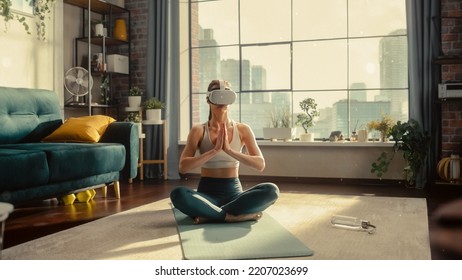 Young Athletic Woman Wearing Virtual Reality Headset In Her Sunny Living Room, Practising Meditation In Modern Futuristic Way. Fit Female Is Focused On Her Wellbeing And Mindfulness.