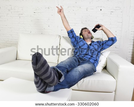 young attractive 20s or 30s man having fun alone lying on home couch listening to music holding mobile phone as microphone using headphones singing passionate happy and crazy
