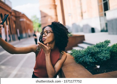 Young attractive African American female in trendy glasses wearing casual clothes blowing kiss while looking at mobile phone camera in street at daytime