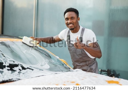 Young attractive African American man washing his yellow luxury car in a self-service car wash station showing thumb up outdoors with cleaning foam and yellow sponge.