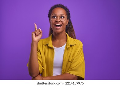 Young attractive African American woman wearing glasses after coming up with idea points finger up and wants to share plan of action dressed in comfortable casual clothes stands on purple background