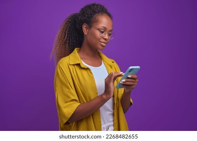 Young attractive African American woman millennial with phone in hands spends all free time on smartphone suffering from gadget addiction and in need of psychological help stands on lilac background
