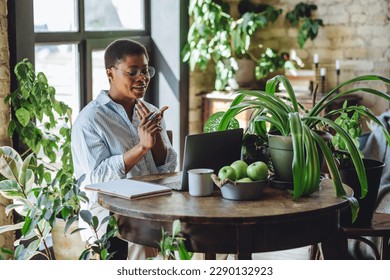 Young attractive African American woman nutritionist is working on laptop in a beautiful green space, home or modern office. Healthy food, apples, water. Video consultation, conference, presentation - Powered by Shutterstock