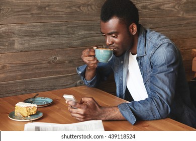 Young and attractive African man in casual dress typing messages in social networks and smiling. He looks so positive and fresh on wooden background, ready to make a sip of fresh brewed coffee. - Powered by Shutterstock