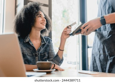 Young attractive african woman paying with credit card on swiping machine in cafe. Female customer client doing cashless payment with debit plastic card in restaurant cafeteria - Powered by Shutterstock