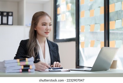 Young attractive Asian business woman with messy desk in office,  sitting at the desk, working at contemporary office and documents. Using calculator. - Shutterstock ID 2232907047