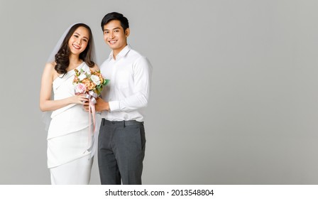 Young attractive Asian couple, man wearing white shirt, woman wearing white dress with wedding veil holding bouquet of flowers together. Concept for pre wedding photography.