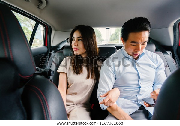 A young and attractive Asian\
couple sit together in the backseat of a car as they are driven to\
their destination. They booked their ride on a ride hailing app.\
