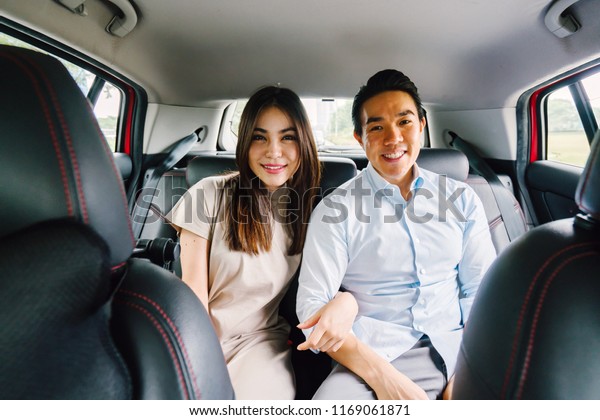 A young and attractive Asian\
couple sit together in the backseat of a car as they are driven to\
their destination. They booked their ride on a ride hailing app.\
