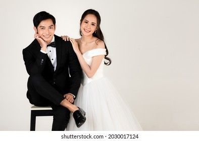 Young Attractive Asian Couple, Soon To Be Bride And Groom, Woman Wearing White Wedding Gown. Man Wearing Black Tuxedo, Sitting Down Together. Concept For Pre Wedding Photography.