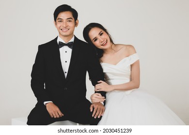 Young attractive Asian couple, soon to be bride and groom, woman wearing white wedding gown. Man wearing black tuxedo, sitting down together. Concept for pre wedding photography.