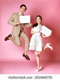 Young attractive Asian couple woman wearing white wedding dress, man wearing beige suit holding signs jumping together against pink background. Concept for pre wedding photography.