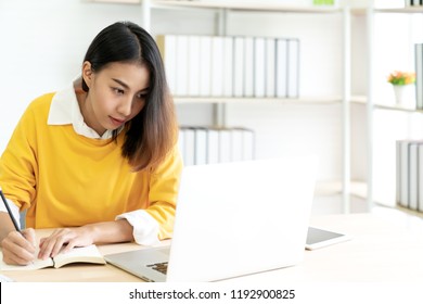 Young attractive asian female student sitting at table looking at laptop writing journal by hand note idea script, diary or sketch design on notebook at home office or library with copyspace concept.