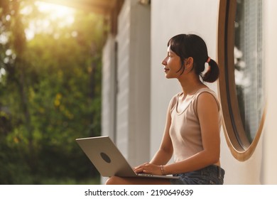 Young attractive Asian freelancer woman sitting on the bench in front of the window house outside working on a laptop computer during summer vacation. Happy Asian teenager student study online. - Shutterstock ID 2190649639