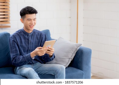 Young Attractive Asian Man Resting Using Browsing Tablet Computer On Sofa At Home, Happy Male Sitting On Couch Relax Reading Digital Gadget At House, Communication And Lifestyle Concept.