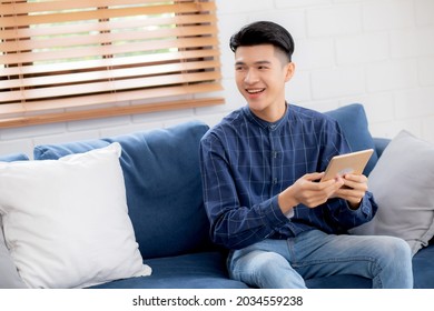 Young Attractive Asian Man Resting Using Browsing Tablet Computer On Sofa At Home, Happy Male Sitting On Couch Relax Reading Digital Gadget At House, Communication And Lifestyle Concept.