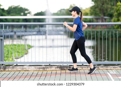 Young Attractive Asian Runner Woman Running At Sidewalk Way In Public Nature City Park With Lake Water View Wearing Blue Or Black Sporty Sportswear With Copy Space In Morning Walk Workout Concept.