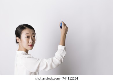 Young attractive asian teacher woman in white shirt writing drawing whiteboard and blue marker   looking at camera over shoulder white background  