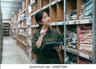 Young Attractive Asian Woman Auditor Staff Work Looking Up Stocktaking Inventory In Warehouse Store. Lady Worker In Clothes Shop Factory Working With Clipboard And Pen. Girl Point At Shelf Counting.