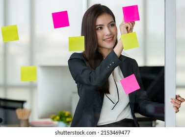 Young Attractive Asian Woman In Black Business Reading And Writing On Post It On Glass Panel In Modern Looking Office With Blurry Windows Background. Concept For Modern Office Lifestyle.