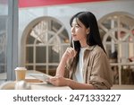 A young attractive Asian woman in casual wear is sitting at a table in a cafe with her digital tablet, gazing at the view outside, daydreaming or thinking about something.