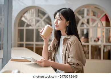 A young attractive Asian woman in casual wear is sitting at a table in a cafe with her digital tablet and sipping coffee while gazing at the view outside and daydreaming. - Powered by Shutterstock