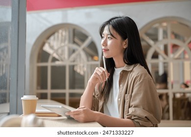 A young attractive Asian woman in casual wear is sitting at a table in a cafe with her digital tablet, gazing at the view outside, daydreaming or thinking about something. - Powered by Shutterstock