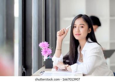 Young attractive asian woman holding pen touching her head and looking at camera while working with business document in co-workspace with her team.