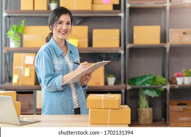 Young Attractive Asian Woman Owner Startup Business Look At Camera Work Happy With Box At Home Prepare Parcel Delivery In Sme Supply Chain, Procurement, Omnichannel Or Coronavirus Quarantine Concept.