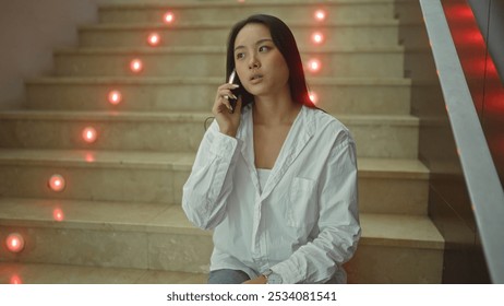 Young attractive asian woman talking on her phone sitting on indoor stairs in a modern corridor with illuminated steps - Powered by Shutterstock