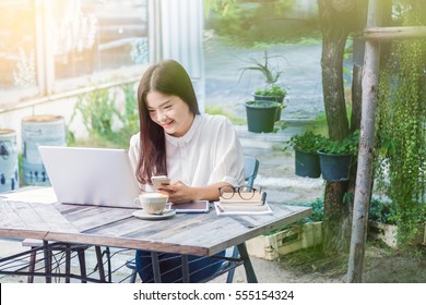 Young Attractive Asian Woman Using Technology On Her Laptop Computer And Her Smart Phone In Outdoor Garden, With Warm Light Flare