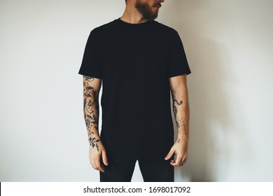 young attractive bearded man with tattoos, dressed in a black blank t-shirt, posing on a white wall background. Empty space for you logo or design.