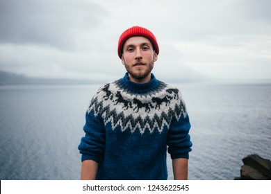 Young attractive bearded millennial man in red fisherman or sailor beanie hat and traditional icelandic ornament blue sweater stands on seaside on cloudy day, serious and tough personality