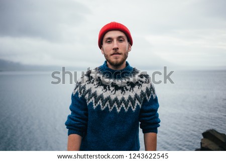 Young attractive bearded millennial man in red fisherman or sailor beanie hat and traditional icelandic ornament blue sweater stands on seaside on cloudy day, serious and tough personality