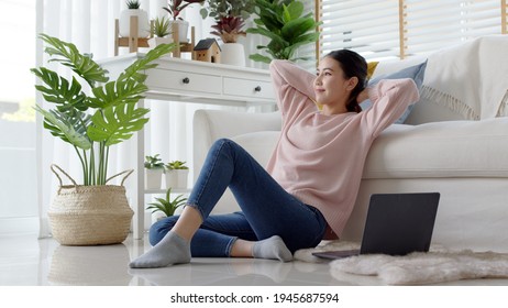 Young attractive beautiful asia female girl or university student sit smile look outside window put arm hand back behind head at sofa couch living room feeling relax comfort at cozy home houseplant.