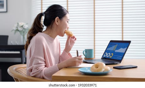 Young Attractive Beautiful Asian Female Hungry Eat Doughnut Take Away Snack Food With Full Mouth Look At Computer Notebook At Home In Busy Work From Home Multitask Unhealthy Meal Lifestyle Concept.