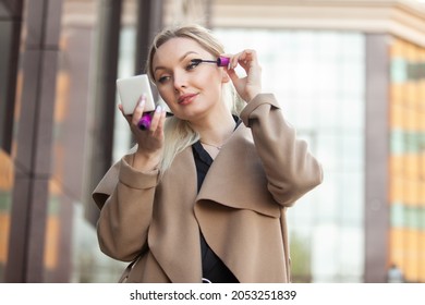 Young attractive beauty woman blonde applying makeup (paint her eyelashes with mascara brush) in city